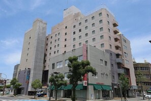 Um edifício de vários andares com uma estrutura vertical destacada à esquerda, uma série de varandas nos andares superiores e uma loja com toldo verde.