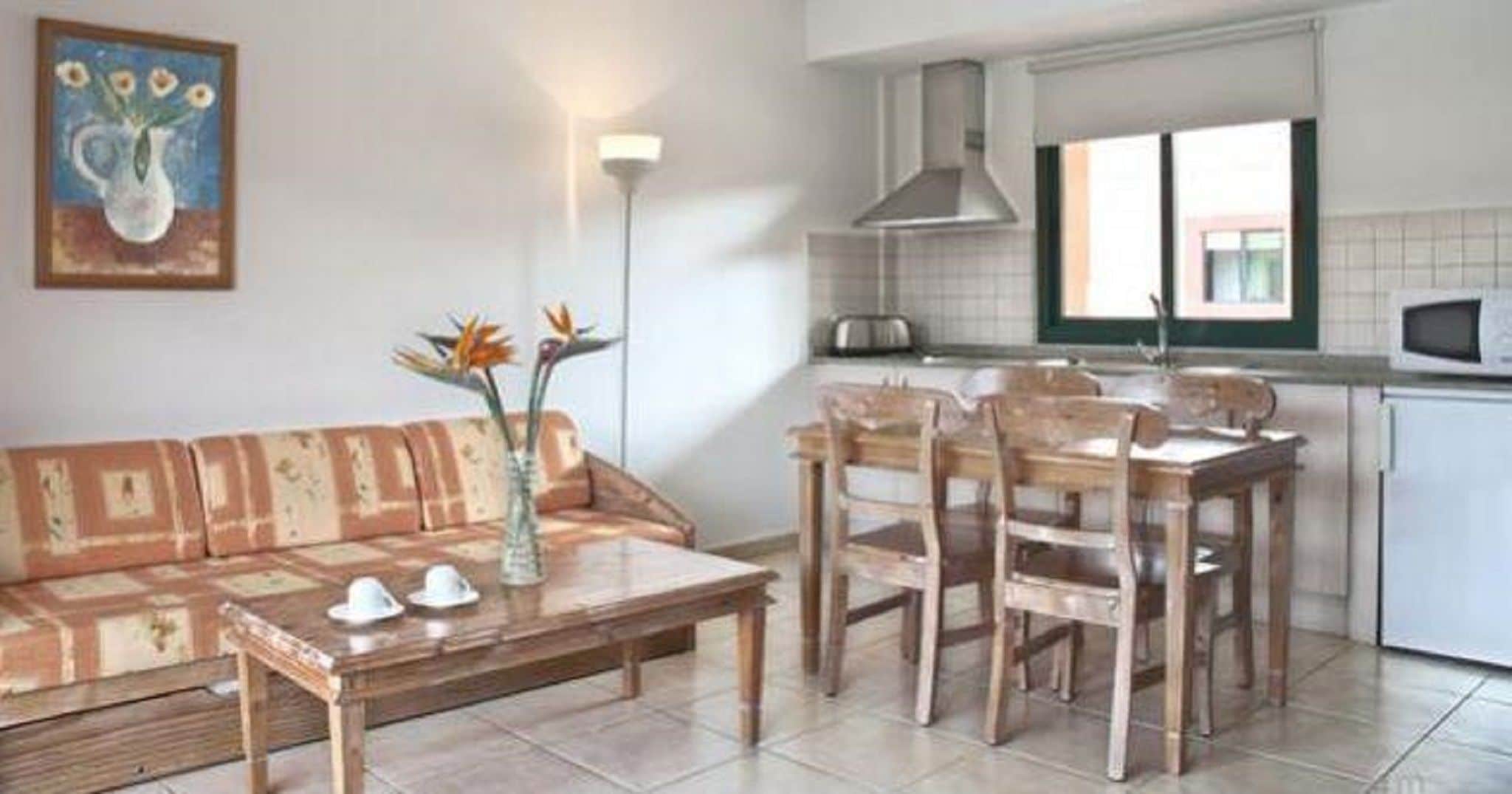 Sala de estar con sofá, mesa de madera y sillas, un jarrón con flores, y una zona de cocina con microondas y armarios.