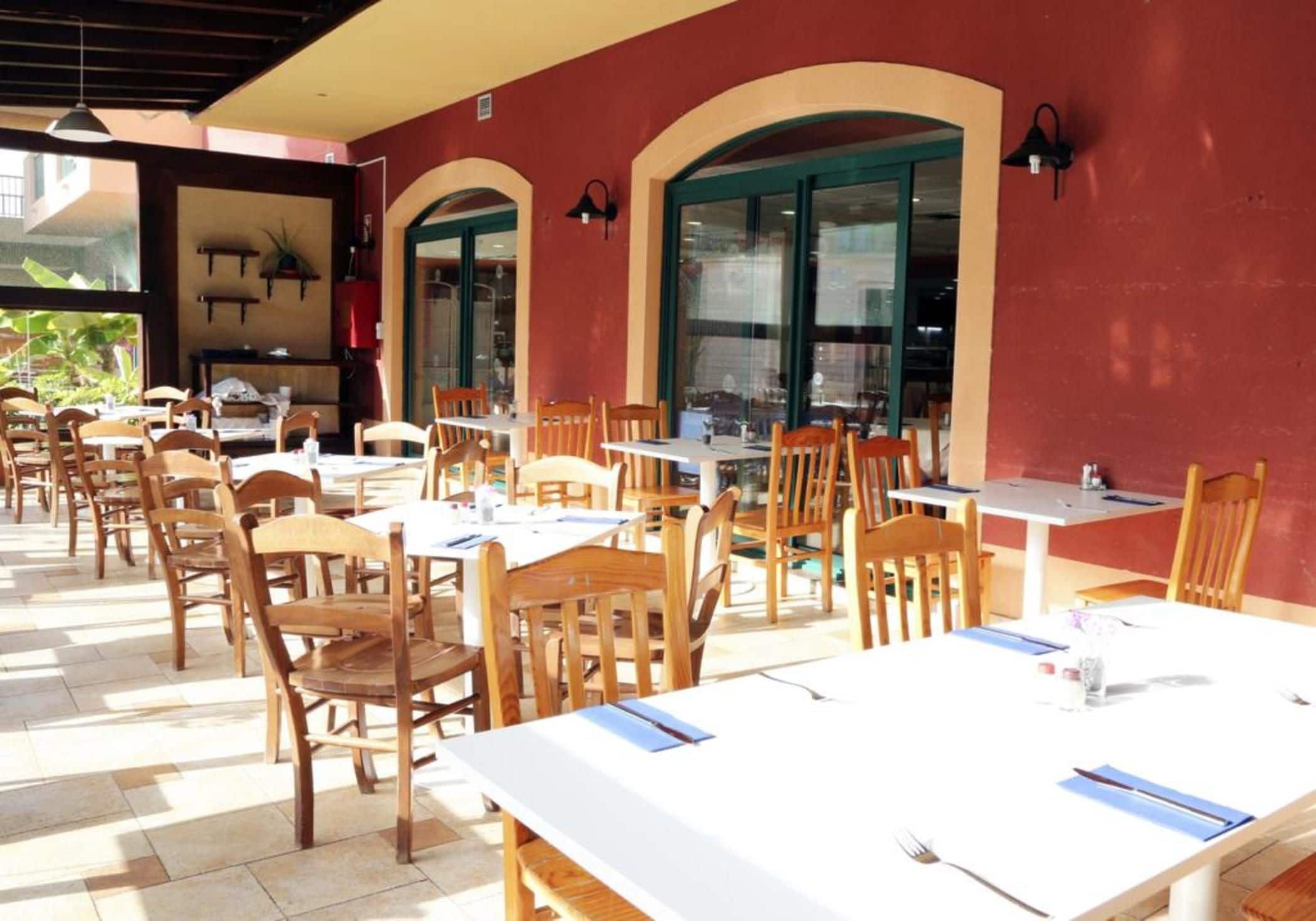 Un espacio de comedor al aire libre con mesas y sillas de madera, ventanas amplias y una pared roja.