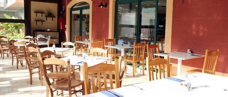 Un espacio de comedor al aire libre con mesas y sillas de madera, ventanas amplias y una pared roja.