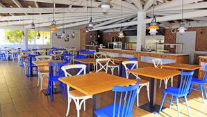 Un vaste espace de salle à manger intérieur, avec des tables en bois et des chaises bleues, une cuisine à l’arrière-plan et un plafond haut avec des poutres apparentes.