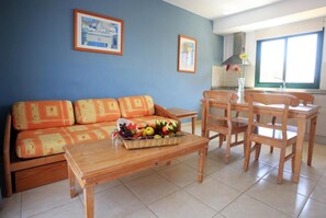 A living room with a sofa, a wooden coffee table with fruit, a dining table with chairs, and two framed pictures on the wall. - O7 Aloe Corralejo (La Oliva)