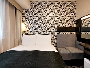 Quarto de hotel com uma cama, uma cadeira, papel de parede estampado e uma mesa de cabeceira.