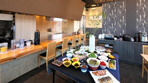 Une cuisine moderne avec un coin repas, comprenant une longue table en bois sur laquelle sont disposés différents plats, une machine à café et un mur en pierre agrémenté d’une plante.
