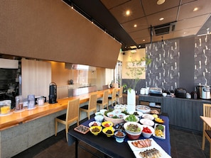 A modern kitchen with a dining area, featuring a long wooden table with various dishes, a coffee machine, and a stone wall with a plant.