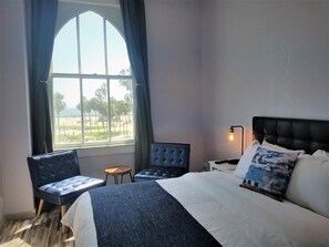 A hotel room with a bed, a chair, a small table, a nightstand with a lamp, and a window with a view of palm trees and a beach. - Venice Beach Cotel (Venice)