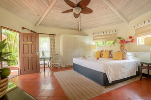 A bedroom with a bed, a ceiling fan, a window with curtains, a door, and a small table with a chair. - East Winds Inn (Gros Islet)