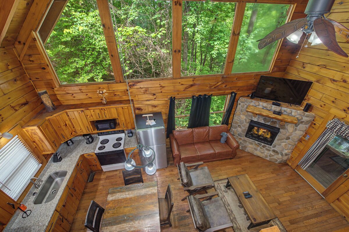 A cabin interior with a kitchen, dining area, fireplace, and large windows overlooking a forest.