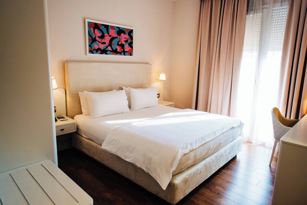 A hotel room with a bed, bedside tables, a lamp, a painting on the wall, and a window with curtains.