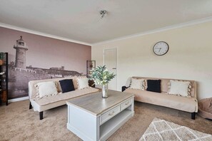Ein Wohnzimmer mit zwei Sofas, einem Couchtisch, einer Wanduhr und einem Wandbild mit einem Leuchtturm.