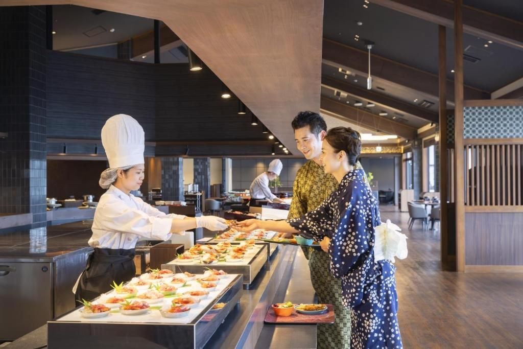 Un'area buffet con diverse postazioni gastronomiche, uno chef che serve cibo e una sala da pranzo con tavoli e sedie.