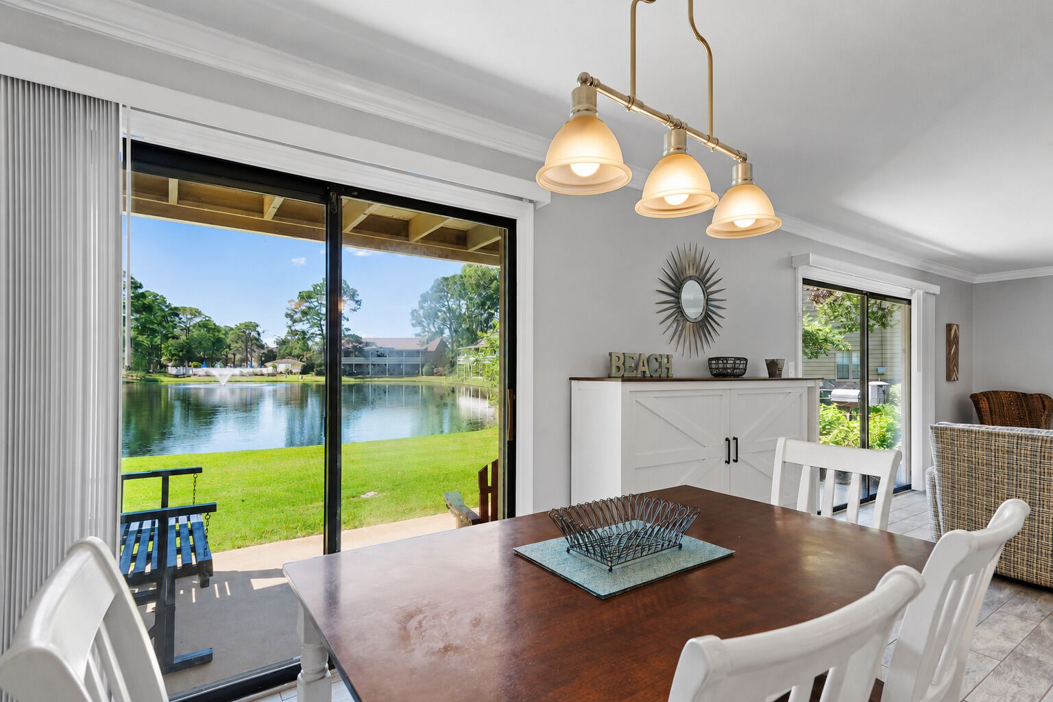 A dining area with a view of a lake, a wooden table, white chairs, and a chandelier.