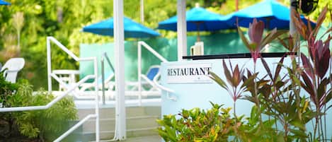 Un espace extérieur abrité, aménagé avec des sièges, et sur lequel est apposée une enseigne indiquant la présence d’un restaurant. L’ensemble est entouré de verdure et agrémenté de parasols bleus.