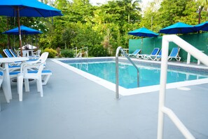 Piscina con sombrillas azules, sillas de descanso blancas y una valla verde.