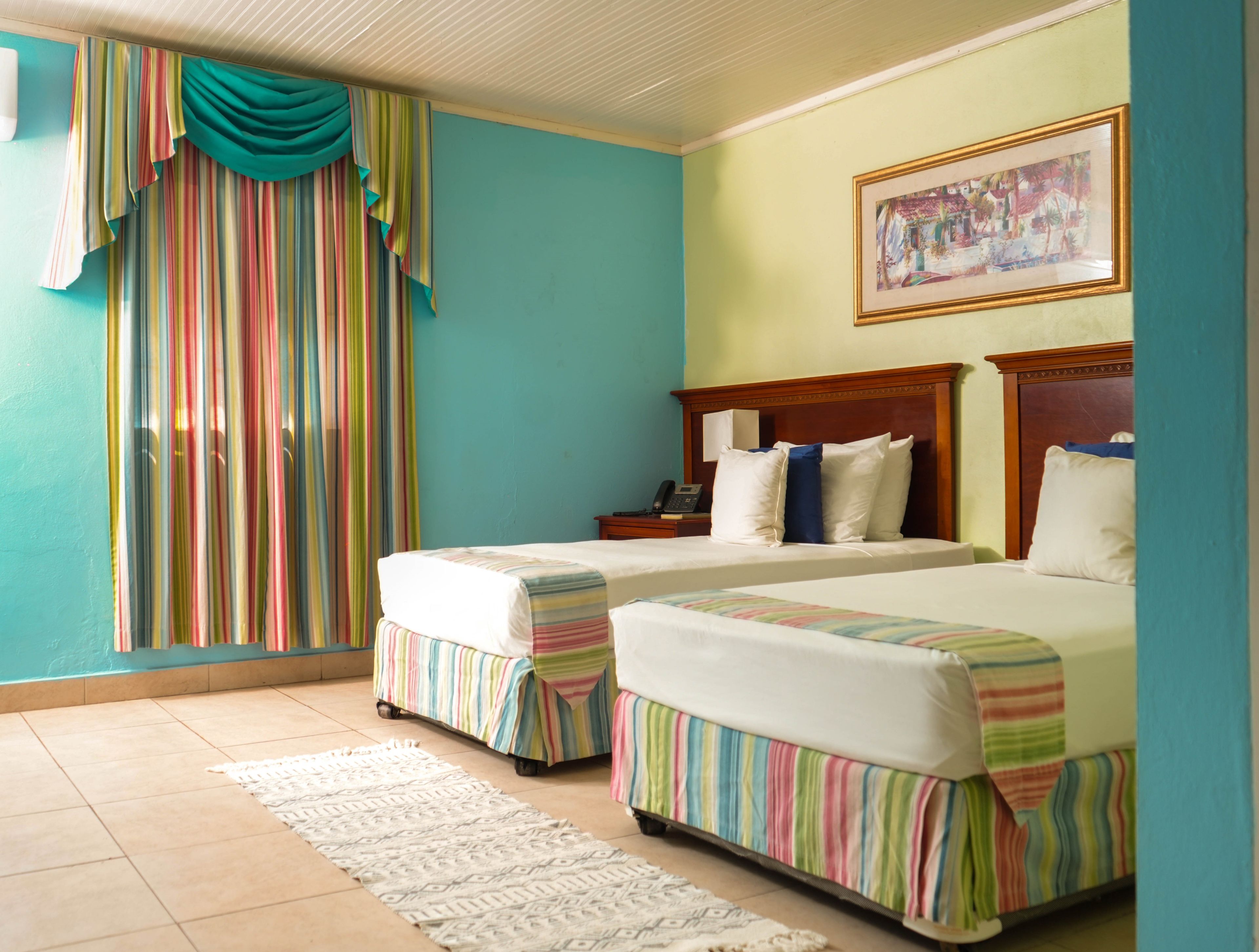 A hotel room with two beds, a large window with curtains, a painting on the wall, and a bedside table with a phone.