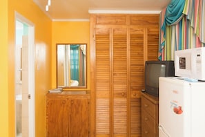 A compact room with a wooden wardrobe, a small cabinet, a television, and a refrigerator. - Vedanta Resort (Worthing)