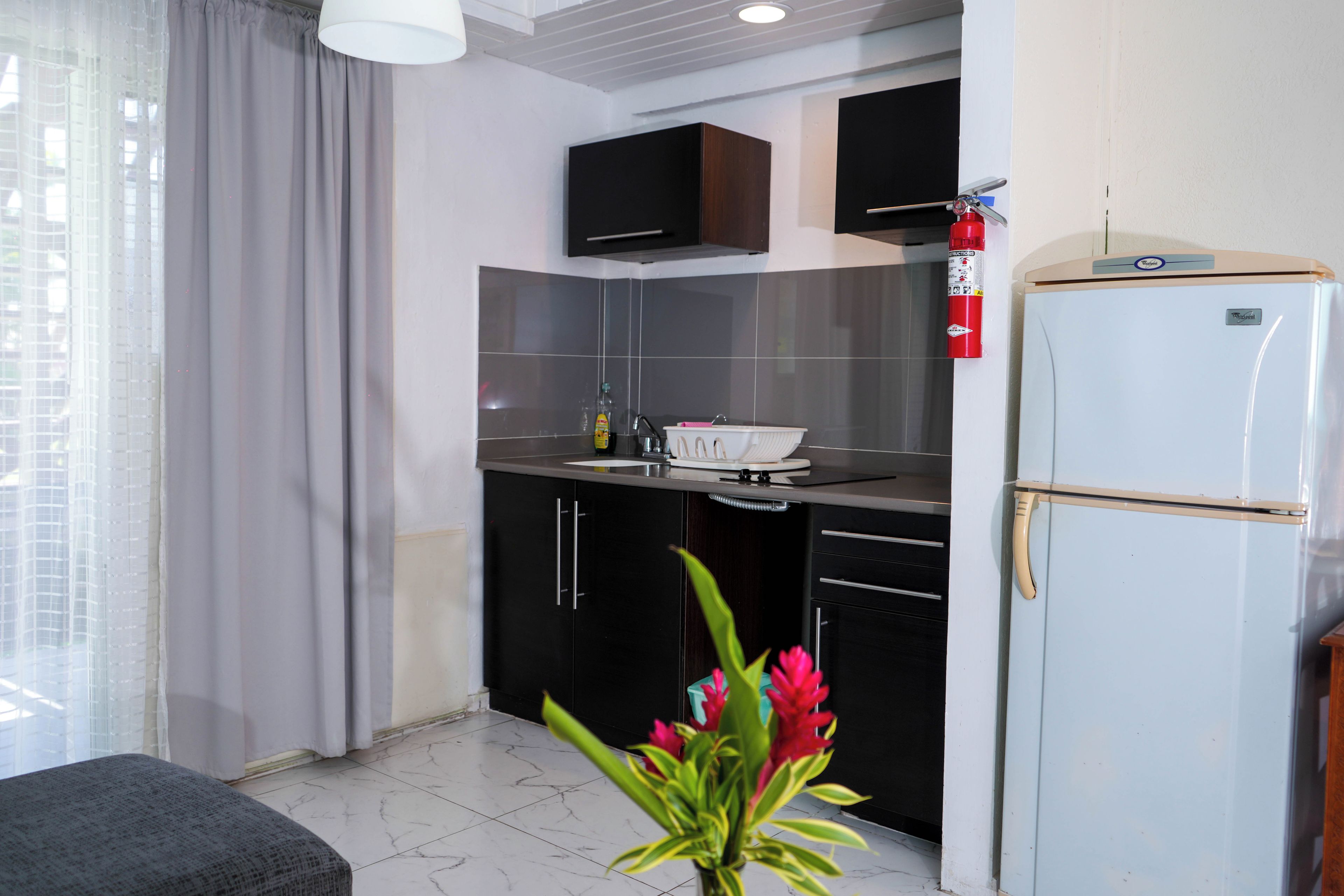 Une cuisine moderne avec des meubles de couleur sombre, un réfrigérateur blanc et une fenêtre ornée de rideaux légers.