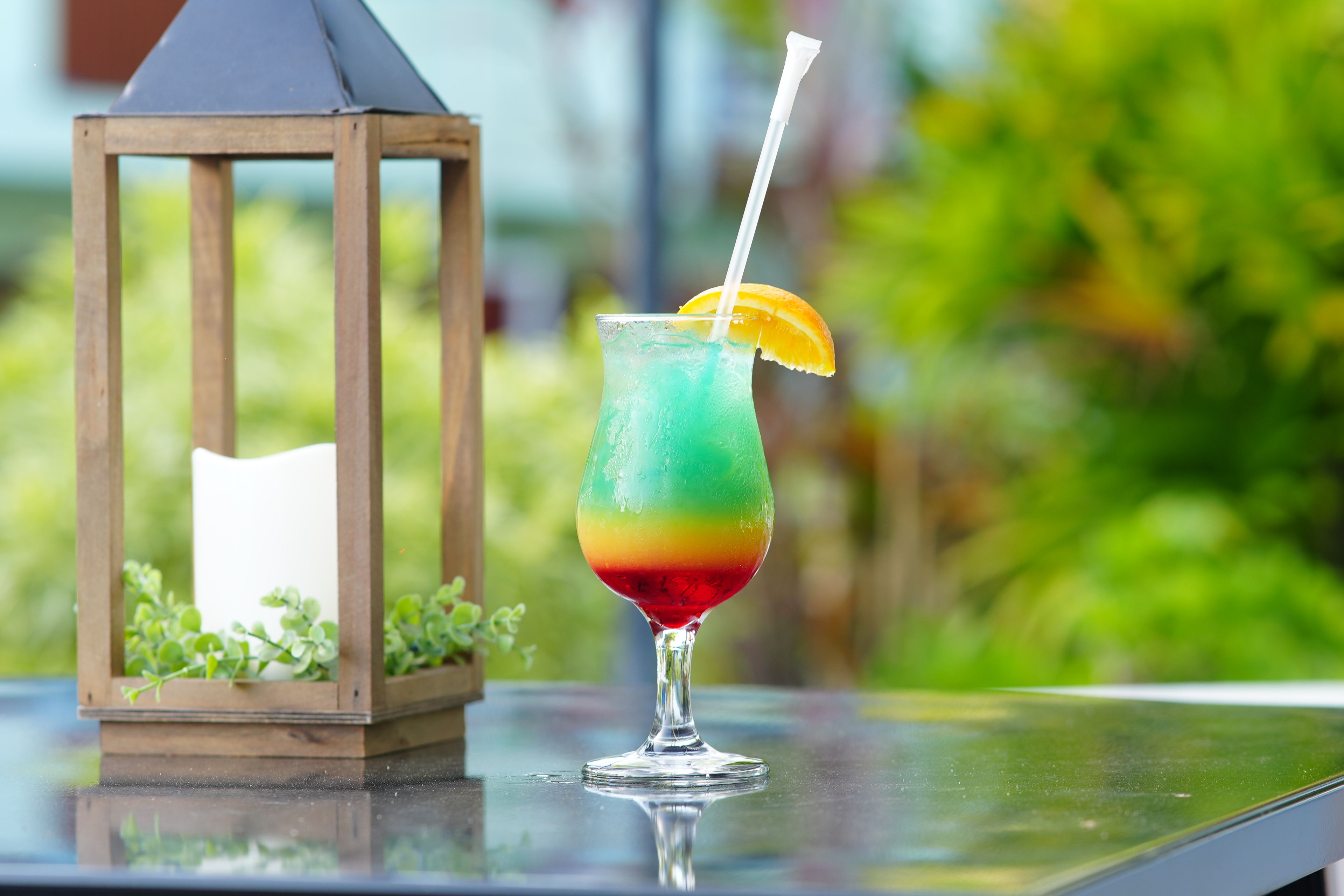 A glass of layered drink with a straw and an orange slice, placed on a reflective surface with a wooden lantern and greenery in the background.