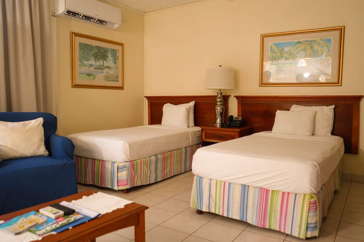 Habitación de hotel con dos camas, una silla azul, una mesita de noche de madera con lámpara y dos cuadros en la pared.