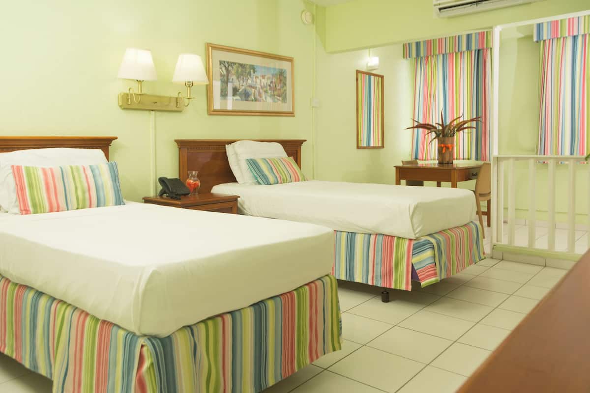 Habitación de hotel con dos camas, una mesita de noche con lámpara, una pequeña mesa con una planta, una ventana con cortinas a rayas y un cuadro colgado en la pared.