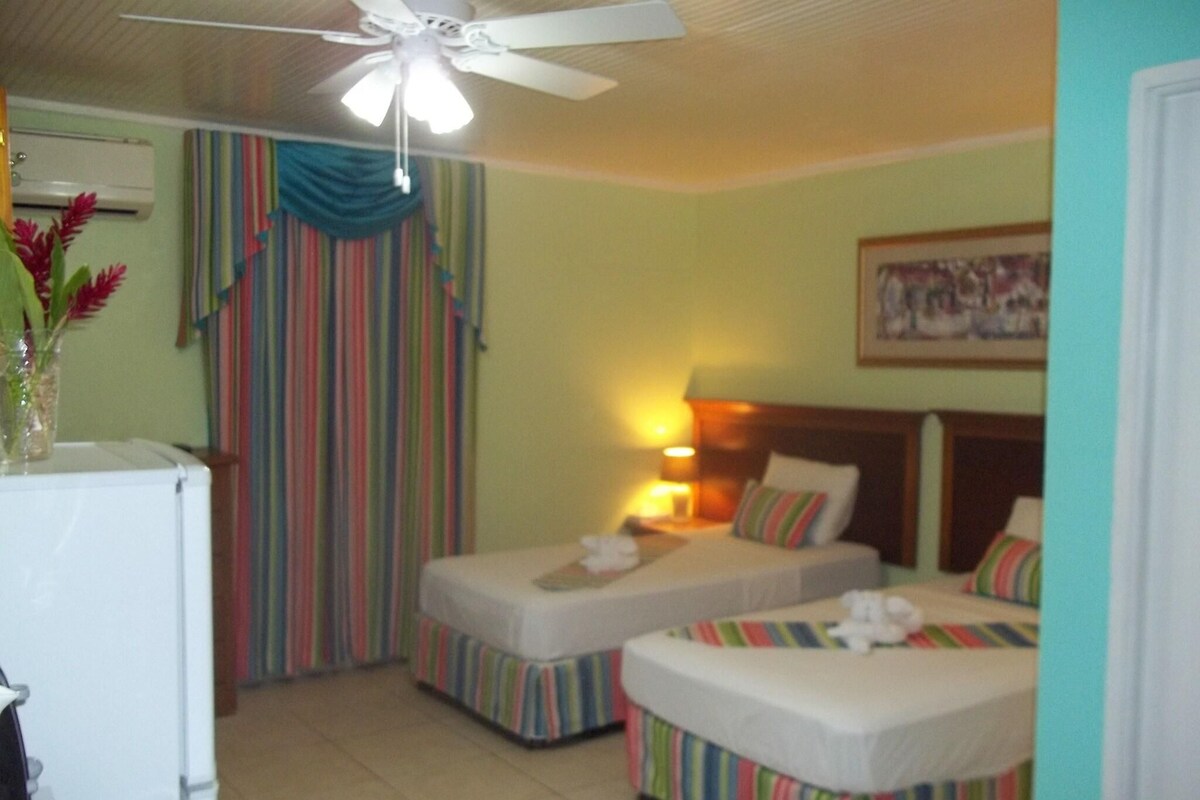 Quarto de hotel com duas camas, um ventilador de teto, uma mesa pequena e um ar condicionado fixado na parede.