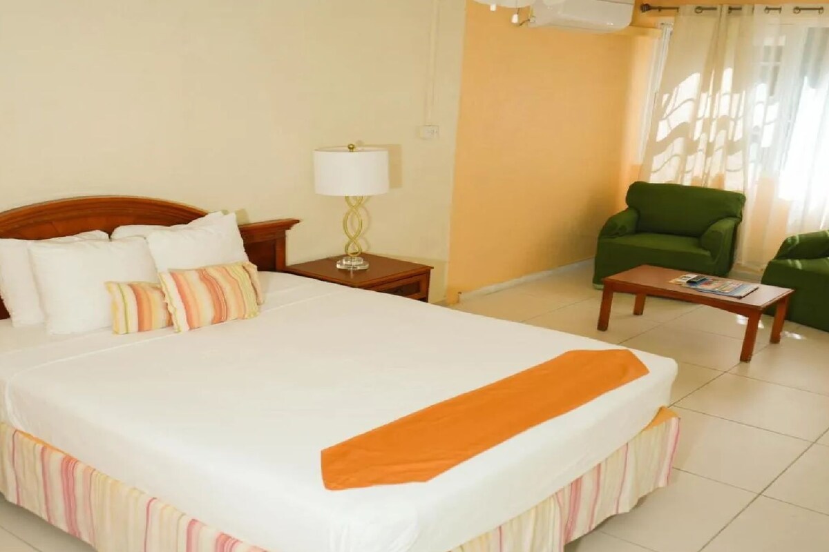 Quarto de hotel com uma cama grande, uma poltrona verde, uma mesa pequena e um abajur.