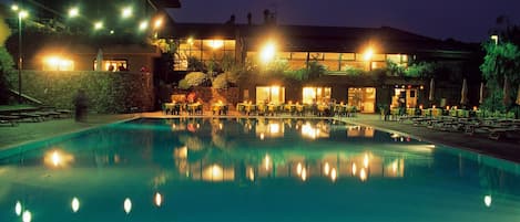 A nighttime swimming pool with surrounding tables and chairs, illuminated by overhead lights.