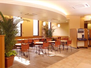 Ein Hotelrestaurant mit runden Tischen und orangefarbenen Stühlen, Zimmerpflanzen und einer Springbrunnen.