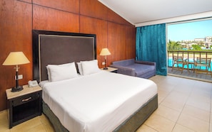 Habitación de hotel con una cama grande, dos mesitas de noche, un sofá y un balcón con vistas a la piscina.