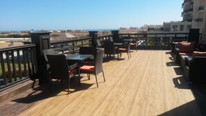 Une terrasse en bois, dotée de mobilier d’extérieur, d’une rambarde et offrant une vue sur la mer et les bâtiments.