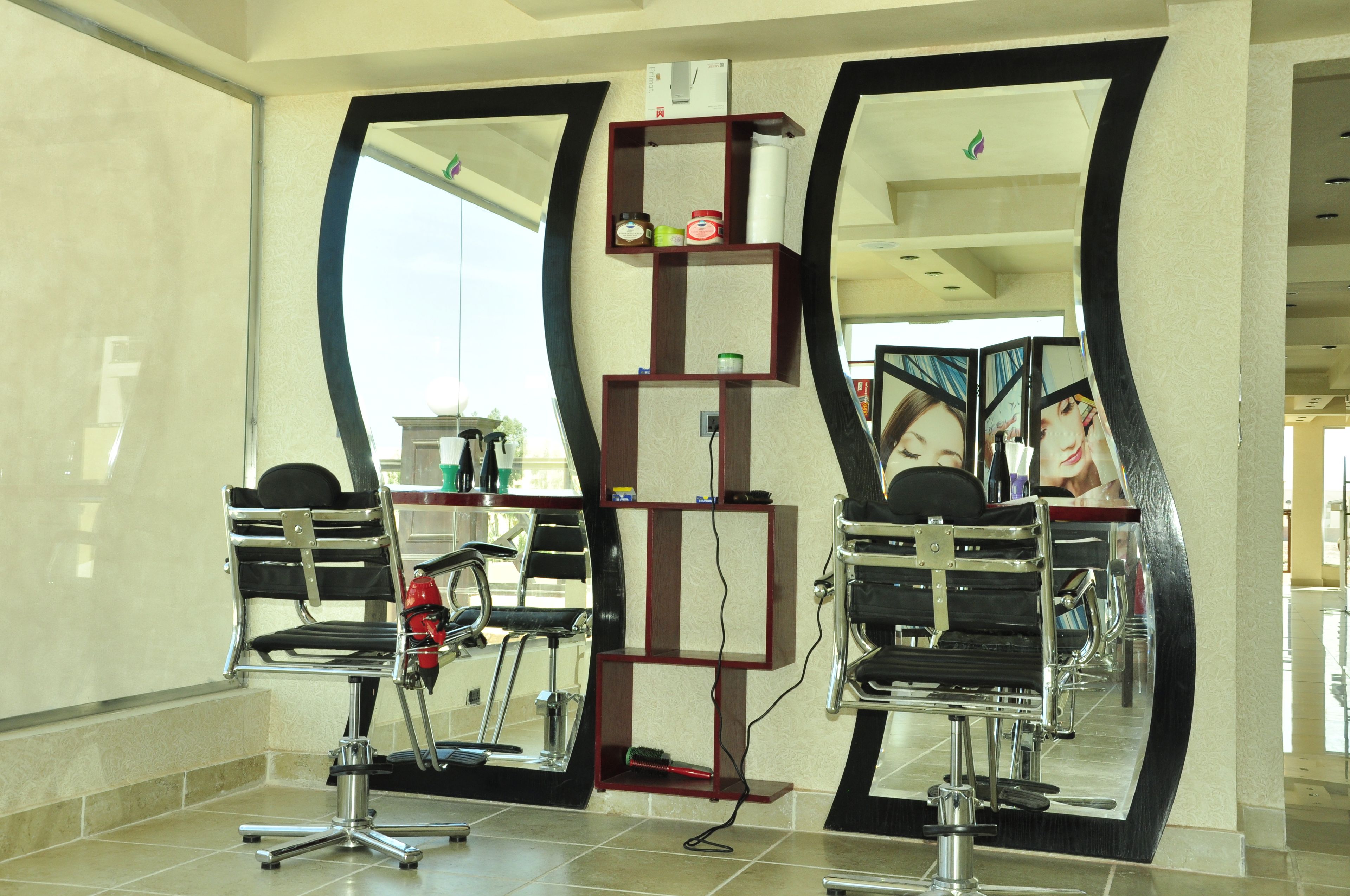 Un salón con dos sillones de barbero con espejo, un estante con productos para el cabello y un espejo grande que refleja el espacio.