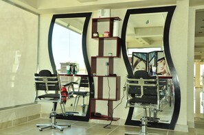 A salon with two mirrored barber chairs, a shelf with hair products, and a large mirror reflecting the room. - Gravity Hotel Aqua Park Hurghada (Hurghada)