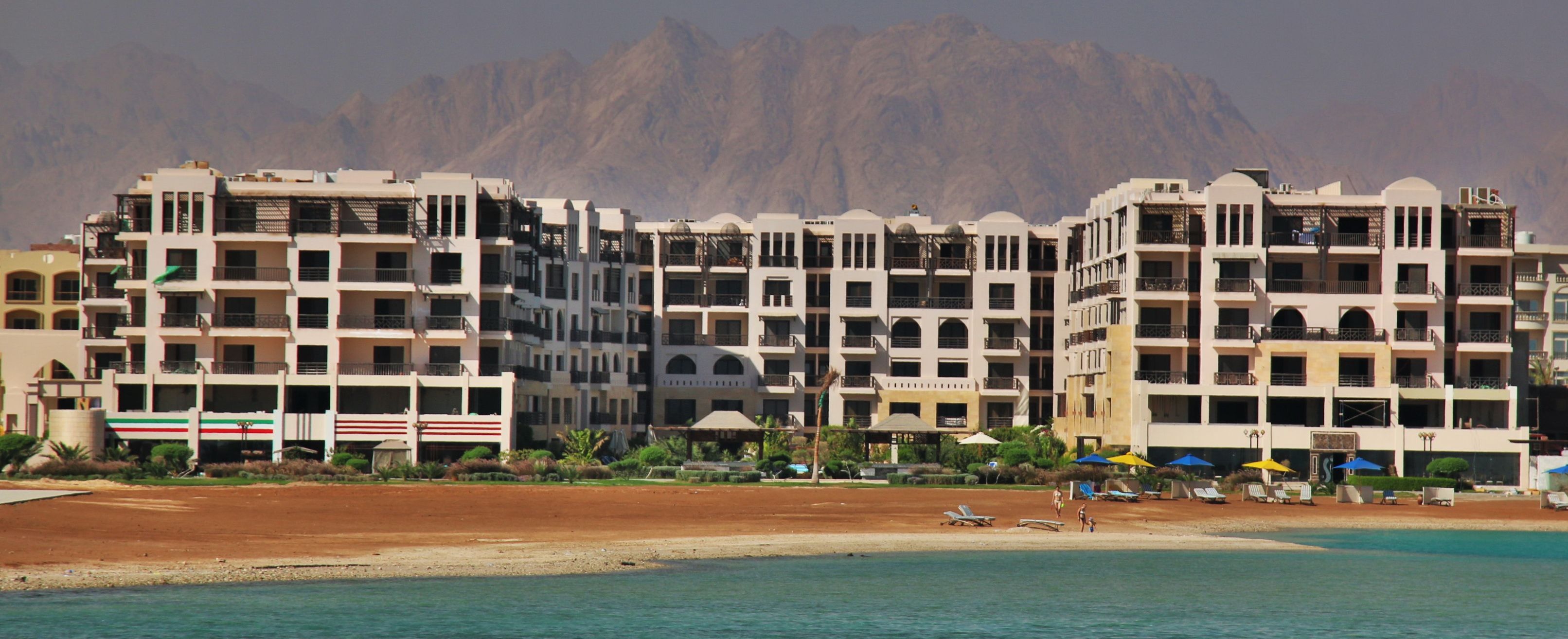 Ein Hotel direkt am Strand mit mehreren Balkonen, einem Sandstrand und einem Berg im Hintergrund.