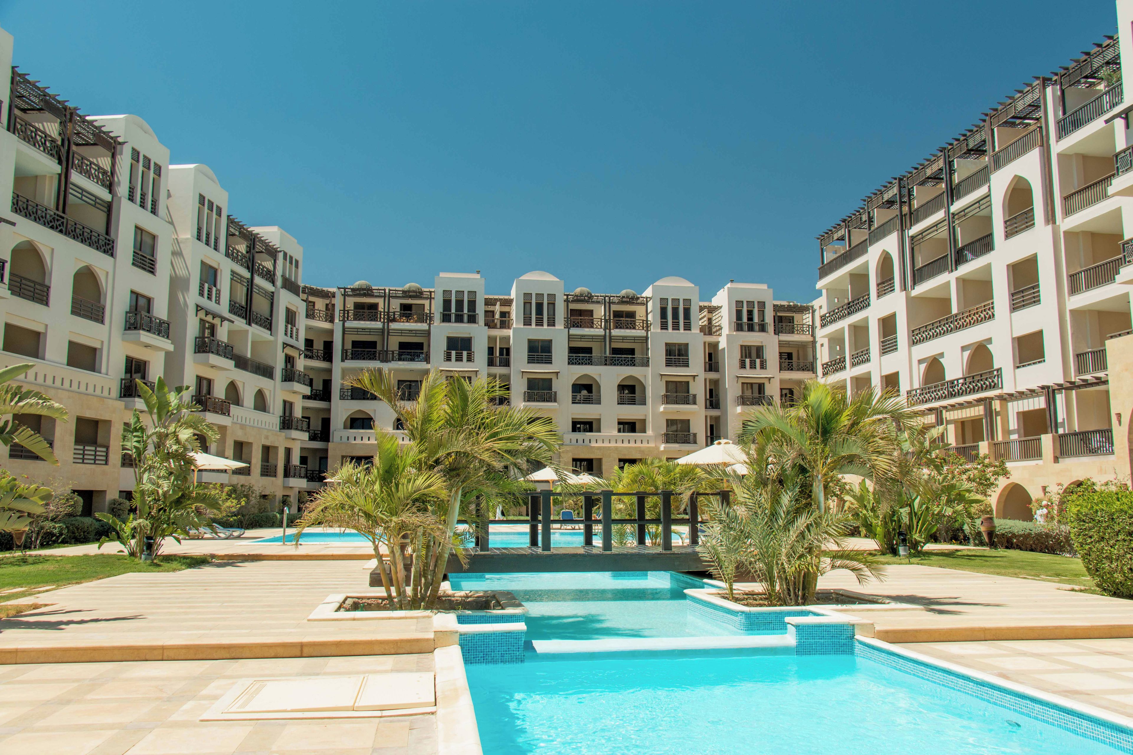 Ein Hotel mit mehreren Etagen, einem zentral gelegenen Schwimmbad und umgebender Bepflanzung.