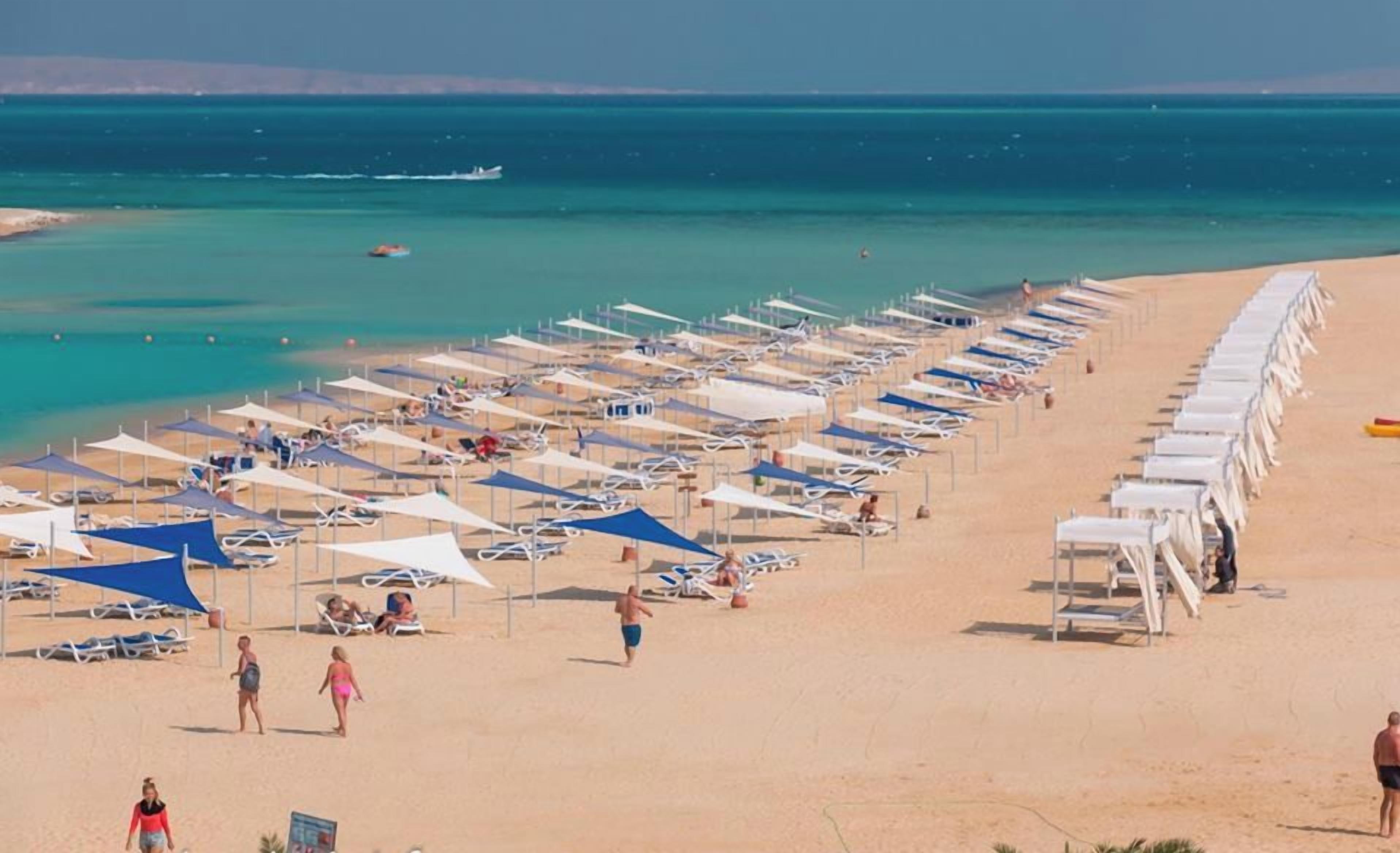Una playa con filas de tumbonas y sombrillas, un mar azul cristalino y una costa distante.