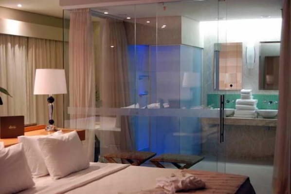 Une chambre d’hôtel comprenant un lit, un bureau, une lampe, une chaise, une petite table et une salle de bain visible à travers une paroi en verre.