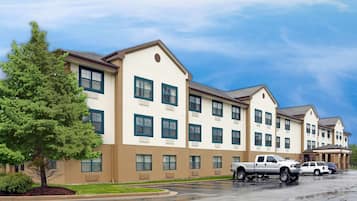 A multi-story hotel with a beige exterior and green window frames, parking area, and a tree.
