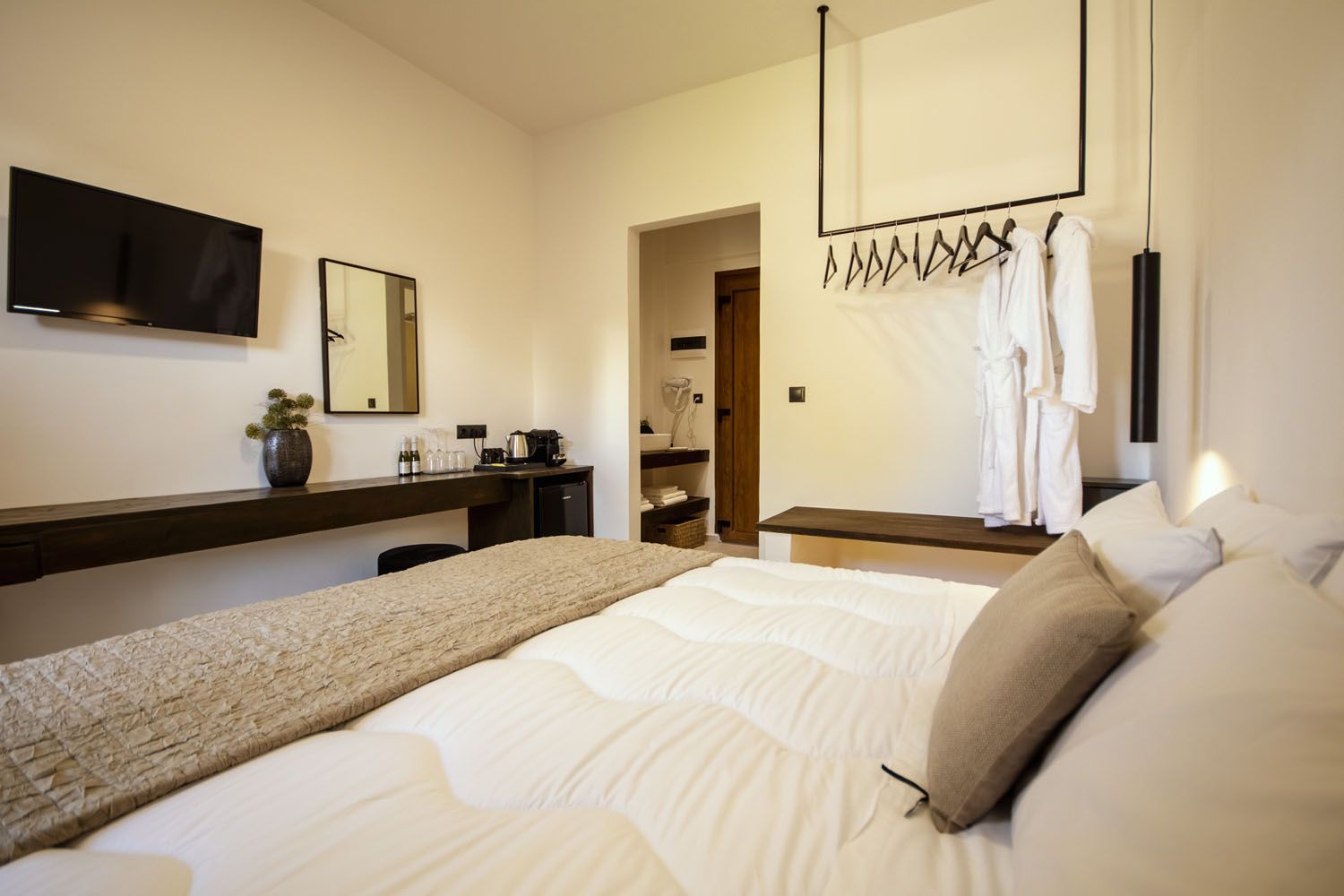 Une chambre d’hôtel avec un grand lit, un bureau doté d’une machine à café, une télévision et un miroir. On y trouve également un porte-peignoir avec des serviettes et une porte donnant accès à la salle de bain.