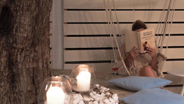 Una persona leyendo un libro en una silla hamaca, con velas y una copa de vino sobre un piso de madera.