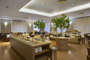 Un espace de restauration moderne dans un hôtel, avec plusieurs buffets, différents types de tables et des plantes décoratives.