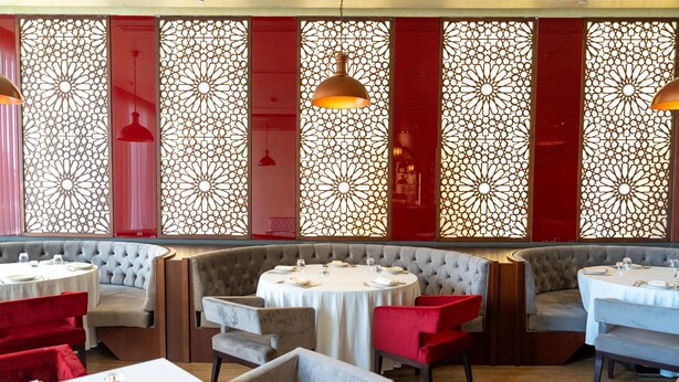 A restaurant with round tables, red and grey upholstered chairs, and decorative red panels. - Levatio Hotel (Muscat)