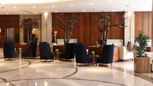 A lobby with a reception desk, dark blue chairs, a large tree sculpture, and a polished marble floor. - Levatio Hotel (Muscat)