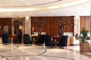 Rebuda amb taulell d'informació, cadires blau fosc, una gran escultura d'arbre i un sòl de marbre polit.