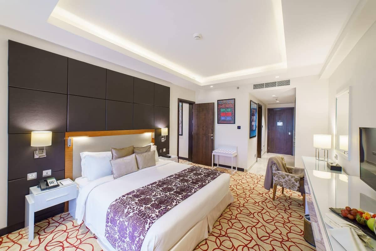 A modern hotel room with a large bed, bedside tables, a desk, a chair, a TV, and a bathroom visible through an open door.