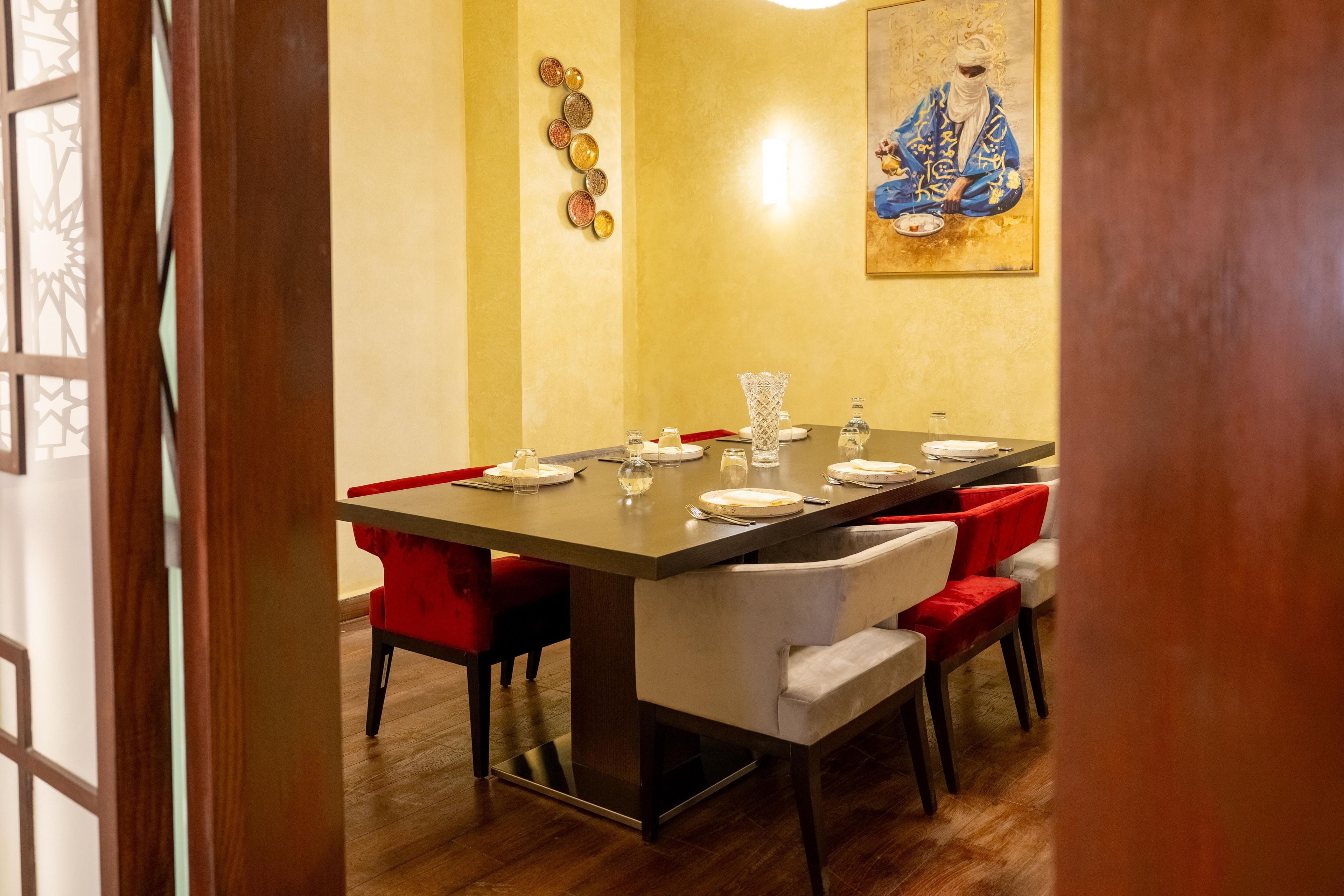 Un comedor con una mesa larga preparada para cuatro personas, sillas tapizadas en rojo, un adorno de pared y un cuadro.