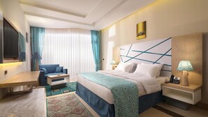 Quarto de hotel com cama, mesas de cabeceira, escrivaninha, cadeira e uma janela grande com cortinas.