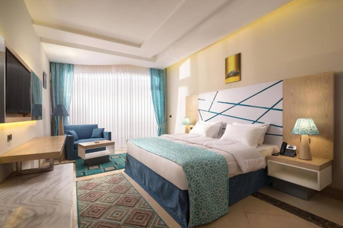 Quarto de hotel com cama, escrivaninha, cadeira, mesa de cabeceira e abajur.
