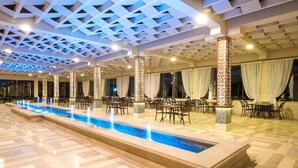 Amplo espaço interno com uma piscina longa, várias mesas e cadeiras e pilares decorativos.