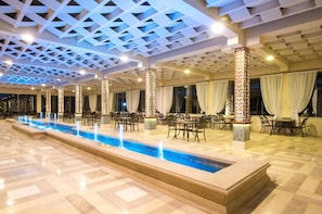 Un amplio espacio interior con una piscina larga, varias mesas y sillas, y pilares decorativos.