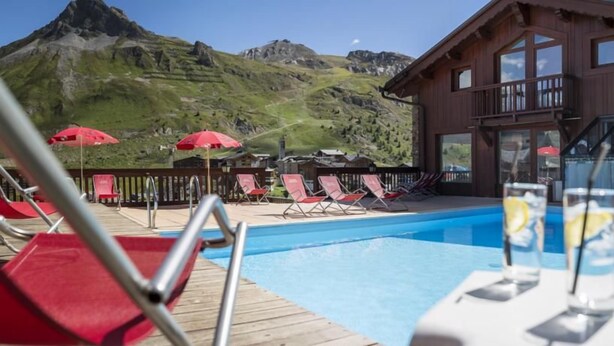 Ett poolområde med solstolar och parasoller, en byggnad i trä med stora fönster och en bergskedja i bakgrunden.